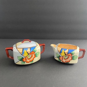 May include: A vintage cream and sugar set. The set features a sugar bowl with a lid and a creamer. Both pieces are decorated with a floral design in orange, red, and green, with geometric shapes in blue, yellow, and gray.