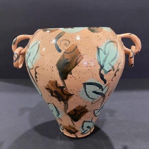 May include: A peach-colored ceramic vase with a speckled finish, featuring two side handles and a hand-painted pattern of light green leaves and dark brown abstract shapes. The vessel has a wide, rounded body that tapers toward the base.