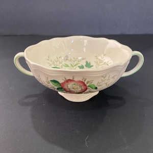 May include: Cream-colored soup bowl with two handles and a floral design. The bowl features a red and white rose, green leaves, and a scalloped rim. The handles are trimmed with green.