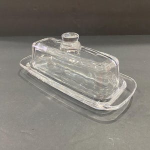 May include: A clear glass butter dish consisting of a rectangular base with slightly raised edges and a matching removable lid. The lid features a small, rounded knob handle on top, and both pieces are made of smooth, transparent glass.