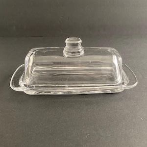 May include: A clear glass butter dish with a rectangular base and a domed lid. The lid has a small, round handle. The dish is designed to hold butter and is made of transparent glass.