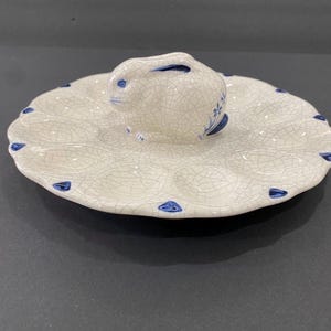 May include: A white ceramic egg serving platter with a bunny figurine in the center. The platter has a scalloped edge with blue accents and a crackle glaze finish. The bunny has blue details.
