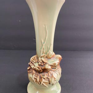 May include: A vintage, light green ceramic vase with a flared top and a detailed, raised relief design. The base features a sculpted floral and bird motif in shades of brown and green. The vase is approximately 8 inches tall.