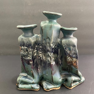 May include: A teal ceramic candle holder with three openings. The piece has a textured, dripped glaze effect with shades of blue, purple, and white. The base is a light brown color.