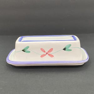 May include: A rectangular ceramic butter dish with a white base and lid. The lid features a hand-painted design of a pink four-petaled flower flanked by two green leaves on thin stems. The edges of the lid and the base are trimmed in purple.