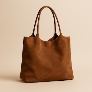 Puede incluir: Un bolso tote de ante marrón con dos asas largas. El bolso tiene forma rectangular con una abertura ancha y un aspecto suave y texturizado. Las asas son del mismo color que el bolso.