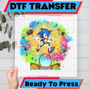 May include: A DTF transfer featuring Sonic the Hedgehog running through a colorful explosion of rings, gems, and other game elements. The text "DTF TRANSFER" and "Ready To Press" are displayed in bold, colorful fonts.