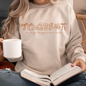 May include: A sand-colored sweatshirt with a fall-themed graphic and the text "in my cozy mom era." The graphic includes a pumpkin, hat, coffee cup, scarf, leaf, and bow. The person is holding a white mug and reading a book.