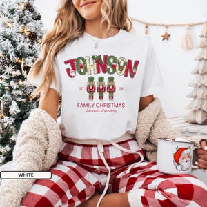 May include: A white t-shirt featuring the red and green text JOHNSON, three nutcracker illustrations, the year 2026, and the text FAMILY CHRISTMAS Jackson, Wyoming. The shirt is worn with red and white plaid pajama pants and a beige cardigan.