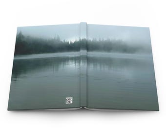 May include: Open journal with a scenic landscape cover. The cover features a misty lake scene with trees on the horizon. The journal is open, revealing the inside pages. The color palette is muted, with shades of gray and green.