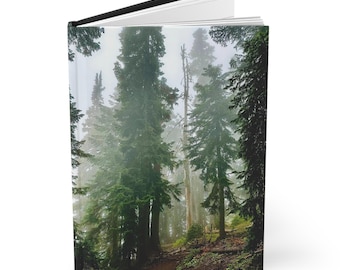 May include: Hardcover journal with a forest scene. The cover features a photograph of tall, green trees in a misty forest. The journal has a black spine and a white interior. The image evokes a sense of nature and tranquility.