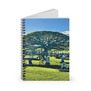 May include: Spiral-bound notebook featuring a vibrant image of a cemetery under a clear blue sky. The image showcases a large tree with a wide canopy, casting shadows over the green grass and numerous headstones.