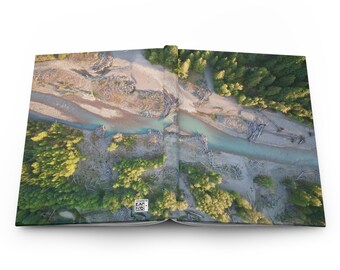 May include: A journal with a landscape image of a river flowing through a forest. The cover shows a bird's-eye view of the river, with green trees and sandy banks. The journal is open, revealing the inside pages.