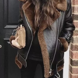 Puede incluir: Chaqueta de cuero negro con forro y ribete de borrego marrón. La chaqueta tiene cierre de cremallera, puños con hebillas y un cuello ancho. Un pequeño bolso de mano beige se sostiene en la mano izquierda. La chaqueta se lleva con pantalones negros.