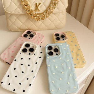 May include: A cream-colored quilted handbag with a gold chain strap and a gold letter "K" is displayed above four phone cases. The phone cases are white, pink, yellow, and blue, each with a polka dot pattern.