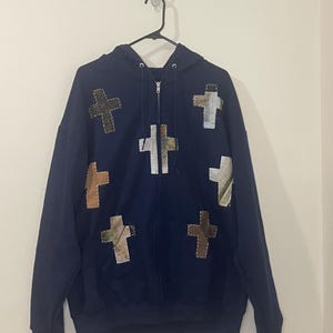 Peut inclure: Un sweat à capuche bleu marine zippé avec plusieurs écussons en forme de croix de différentes couleurs et textures. Les croix sont cousues sur le devant. Le sweat est suspendu à un cintre noir.