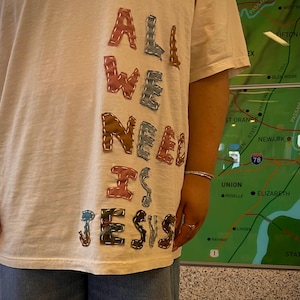 以下が含まれることがあります： クリーム色のTシャツ。カラフルなパッチワーク風の文字で「ALL WE NEED IS JESUS」と書かれています。クルーネックと半袖です。青いジーンズを履いている人がいます。