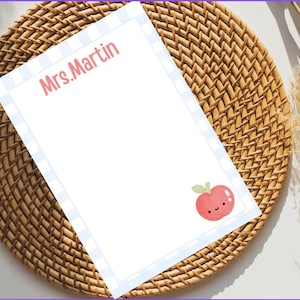 Peut inclure: Bloc-notes blanc avec une bordure à carreaux bleus et blancs et le nom "Mrs. Martin" en rose. Une pomme de dessin animé avec un visage souriant est dans le coin. Le bloc-notes est sur un dessous de plat en osier tressé.