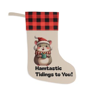 Op de afbeelding: Een kerstsok met een beige body en een rood en zwart geblokte manchet. De sok heeft een cartoon hamster met een kerstmuts en sjaal, die een mok vasthoudt. De tekst luidt "Hamtastic Tidings to You!"