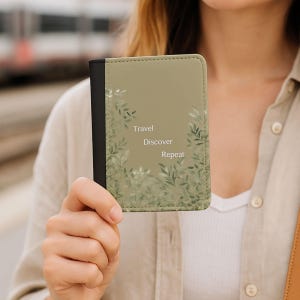 May include: A passport holder with a sage green cover and a black spine. The front features the words "Travel, Discover, Repeat" with a leafy design in shades of green. The holder is made of a smooth material.