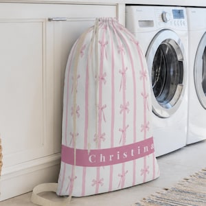 May include: A white drawstring laundry bag featuring vertical light pink stripes and a repeating pattern of small pink bows. A horizontal pink band across the lower section displays the name Christina in white, sans-serif capital letters.