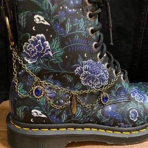 Puede incluir: Bota de combate negra con un estampado floral en azul, verde y blanco. La bota presenta una cadena dorada con abalorios de piedras preciosas azules y un colgante de pluma. Cordones negros y ojales metálicos.
