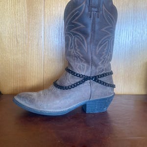 May include: A brown leather cowboy boot with decorative stitching and a black chain ankle accessory. The boot has a pointed toe and a stacked heel. The boot is displayed on a brown surface.