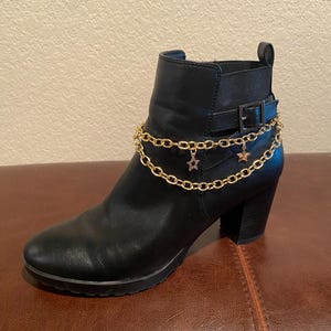 May include: Black ankle boot with a gold chain ankle bracelet featuring star charms. The boot has a block heel, a buckle detail, and a platform sole. The boot is made of a smooth, leather-like material.