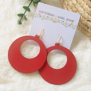 May include: A pair of large, red, circular wooden earrings with gold-colored hooks. The earrings are displayed on a white card with the text "SOUTHERN GIRL ROSE" and a floral design. The background is a soft, white, textured surface.