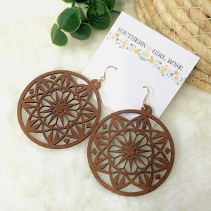 May include: Two pairs of brown wooden earrings with a circular, openwork design featuring a starburst pattern. The earrings have gold-colored hooks and are displayed on a white card with the text "SOUTHERN GIRL ROSE".