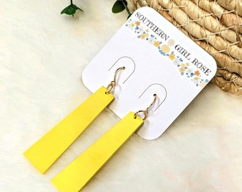 May include: A pair of bright yellow rectangular earrings with gold-colored hooks. The earrings are displayed on a white card with the text "SOUTHERN GIRL ROSE" and floral accents. The background is a soft, white, textured surface.