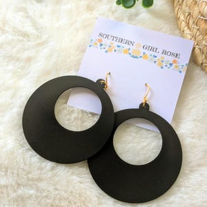 May include: A pair of large, black, circular earrings with gold-colored hooks. The earrings are displayed on a white card with the text "SOUTHERN GIRL ROSE" and a floral design. The background is a fluffy, white surface.