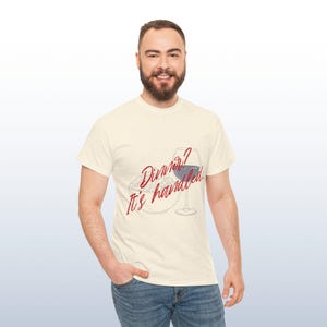 May include: A cream-coloured t-shirt featuring the text "Dinner? It's handled" in red script, alongside a wine glass and a plate of food. The t-shirt is worn by a person.