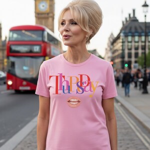 以下が含まれることがあります： カラフルな文字で「Tipsy」と唇のグラフィックがプリントされたピンク色のTシャツ。ロンドンの赤い2階建てバスとビッグ・ベンを背景に着用されています。