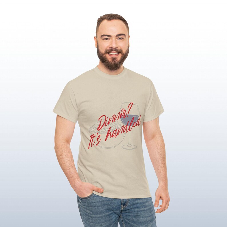 May include: Beige t-shirt featuring the text "Dinner? It's handled" in red script. The graphic includes a plate of food and a wine glass with a blue liquid. The person is wearing blue jeans.
