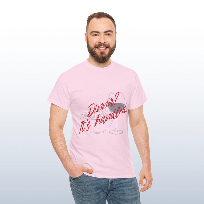 May include: A pale pink t-shirt featuring the text "Dinner? It's handled." in red script. The graphic includes a wine glass and a plate of food. The shirt is worn by a person.