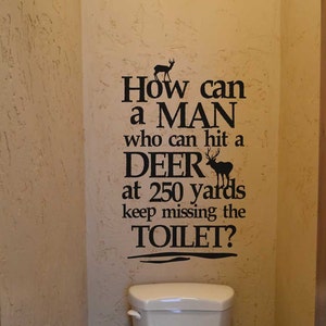 May include: A cream-colored toilet is centered in a bathroom, with a humorous quote in black lettering on the wall: "How can a MAN who can hit a DEER at 250 yards keep missing the TOILET?" The walls are textured, and a wooden door is visible.