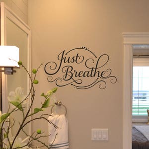 Pode incluir: Interior de casa de banho com um autocolante de parede com a frase "Just Breathe" em escrita cursiva. As paredes são bege neutro. Um espelho, um aplique de parede e um arranjo floral com flores brancas também são visíveis.