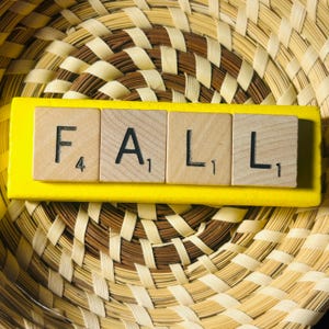 Può includere: Un elemento decorativo con la parola "FALL" scritta con tessere di Scrabble su una base giallo brillante. Le tessere poggiano su un cestino intrecciato con una palette di colori naturali, creando un tema autunnale.