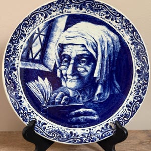 Puede incluir: Plato decorativo azul y blanco con una ilustración detallada de una persona mayor leyendo un libro. El plato tiene un borde floral detallado y se exhibe en un soporte negro. La obra de arte recuerda a la cerámica de Delft.