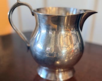 May include: A polished silver-toned metal creamer pitcher featuring a rounded body, a flared pedestal base, a curved handle, and a pointed pouring spout. The reflective surface shows light highlights and subtle signs of use across its exterior.
