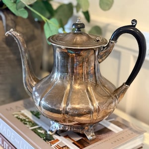 May include: A silver-colored teapot with a black handle and a decorative lid. The teapot has a ribbed design and sits on three small feet. The teapot is placed on top of a stack of books.