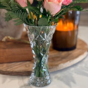 May include: A clear, cut-glass vase filled with pink roses and green foliage. The vase has a diamond pattern and is set on a white surface. A wooden tray and a candle in a brown glass holder are in the background.