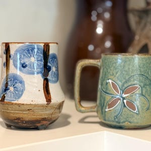 Puede incluir: Dos tazas de cerámica. Una es blanca con diseños florales azules y detalles marrones. La otra es verde con un diseño floral marrón y blanco. Ambas tazas tienen asas y están sobre una superficie blanca.