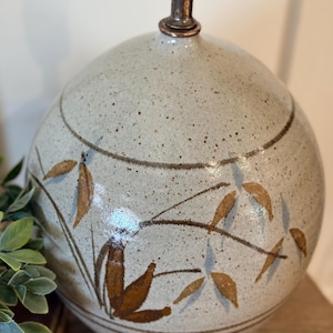 May include: A spherical ceramic table lamp base with a speckled light gray glaze. The surface features hand-painted brown and muted blue leaf motifs arranged in horizontal bands. A metallic bronze-toned neck extends from the top center.