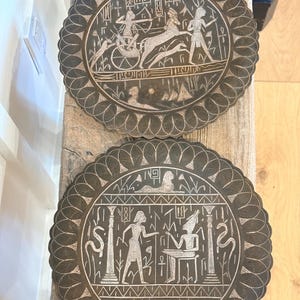 Könnte beinhalten: Zwei dekorative, runde, bronzefarbene Teller mit aufwendigen Gravuren im ägyptischen Stil. Jeder Teller hat einen gewellten Rand und detaillierte Szenen von Figuren, Streitwagen und Hieroglyphen. Die Teller werden auf einer Holzoberfläche präsentiert.