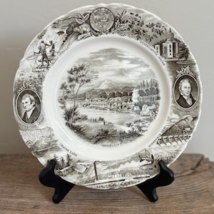 May include: A decorative plate with a black and white landscape design, featuring historical figures and landmarks. The plate includes text such as "Crater Lake" and "Oregon City". It is displayed on a black stand, showcasing its intricate details.