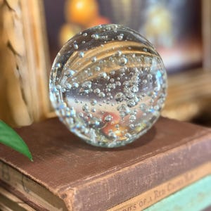 May include: A clear glass paperweight with numerous air bubbles, resting on a stack of antique books. The paperweight is spherical and reflects light, creating a shimmering effect. The books have worn, brown covers.
