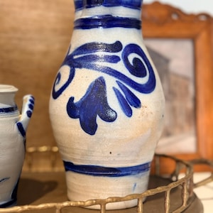 May include: A vintage, off-white ceramic pitcher with blue floral designs and a spout. The pitcher features a blue band around the top and bottom. It is displayed on a gold-colored tray.