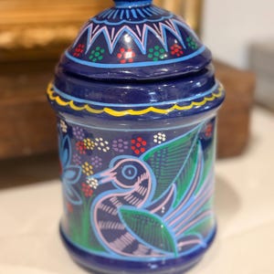 May include: A cobalt blue ceramic jar with a lid. The jar is decorated with a bird and floral design in shades of pink, green, and blue. The lid has a pink flower on top and a geometric pattern.